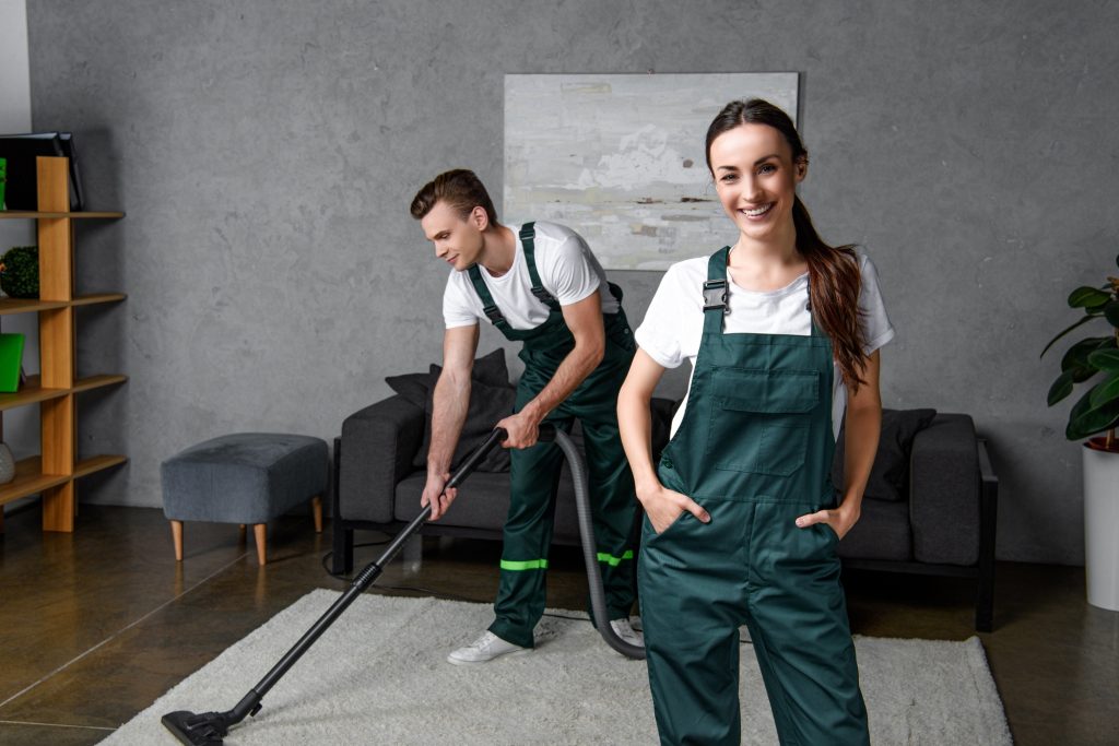 young cleaning company workers using vacuum cleaner and smiling at camera